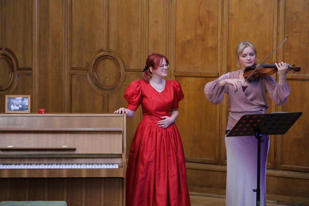 Dwie młode kobiety w czerwonej i liliowej sukni. Jedna z nich trzyma rękę na pianinie, druga gra na skrzypcach.