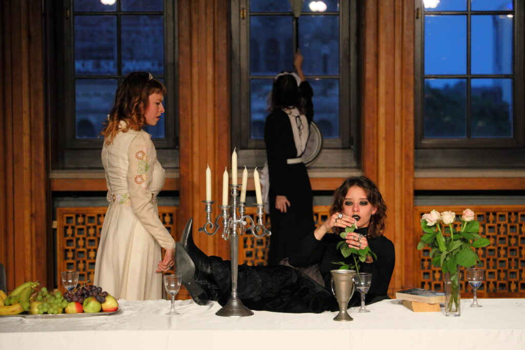 Młoda kobieta w czerni trzyma nogi na stole i obrywa płatki białej róży. Na stole wazon z różami, świecznik, puchar i taca z owocami. Obok bokiem stoi młoda kobieta w białej sukni. W tle odwrócona plecami osoba czyści okno.