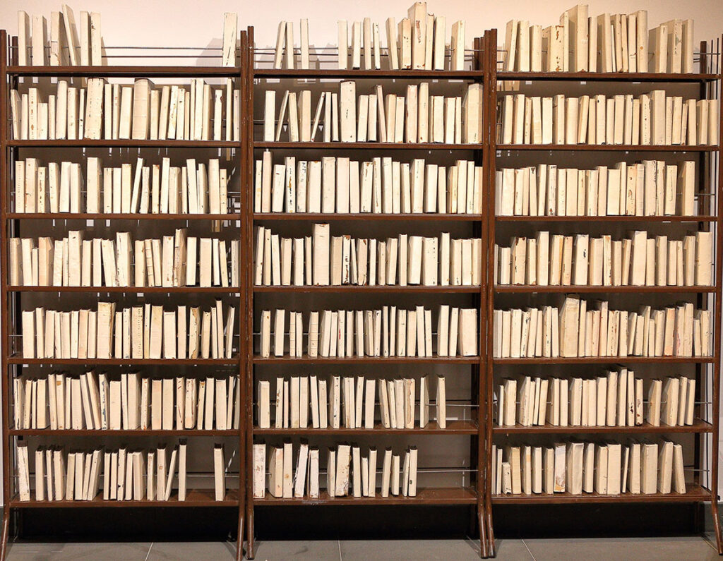 Na zdjęciu widzimy trzy brązowe, metalowe regały, które w całości wypełnione są różnymi książkami bez okładek. Wszystkie książki wyglądają podobnie, różnią się jedynie wielkością i wysokością.