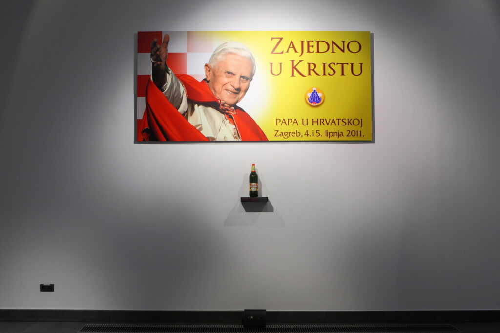 Na białej ścianie zwieszony jest kolorowy, czerwono-żółto-biały baner z wizerunkiem papieża Benedykta XVI i informacją o dacie pielgrzymki Ojca Świętego do Chorwacji. Pod wydrukiem widzimy małą półeczkę, na której stoi butelka piwa lub wina.