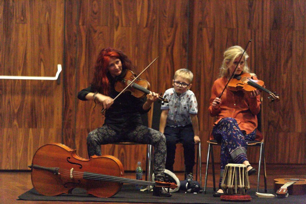Fotografia przedstawia dwie artystki grające na skrzypcach oraz chłopca. Wszyscy siedzą na krzesłach, na tle pokrytej boazerią ściany. Kobieta po lewej stronie ma długie, bujne, kasztanowo-czerwone włosy. Ubrana jest w czarną bluzkę i ciemne spodnie w jasnopopielate wzory. Trzymanym w prawej dłoni smyczkiem gra na skrzypcach. Na podłodze przed artystką leży wiolonczela. Druga kobieta ma długie blond włosy. Ubrana jest w pomarańczową koszulę i niebieskie spodnie w kolorowe wzory. Trzymany pod brodą instrument strunowy podpiera lewą dłonią. Smyczek, który trzyma w prawej ręce, uniesiony jest w górę, w każdej chwili gotowy do użycia. Pomiędzy skrzypaczkami przysiadł mały chłopiec. Ma krótkie blond włosy i okulary. Ubrany jest w jasnobłękitną koszulę z krótkimi rękawami i ciemne spodnie.