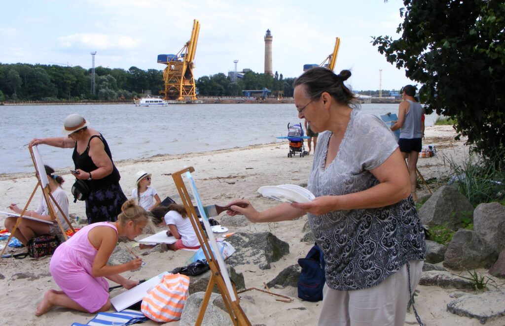 Plaża w Świnoujściu. Dzieci, młodzież i dorośli malują pejzaż, trzymając prace na kolanach lub przenośnych sztalugach. W oddali widać port.