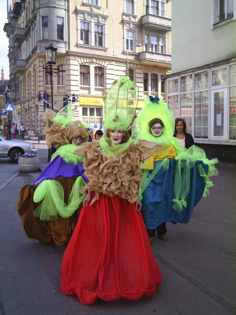 Performance na ulicy miasta – trzy kobiety w dużych, obszernych kostiumach, wykonanych z tkanin, papieru i drutu.