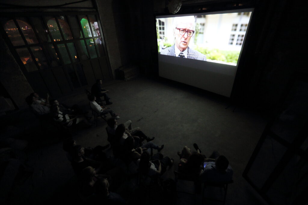Duże, ciemne pomieszczenie, betonowa podłoga. Grupka widzów siedzi na krzesłach dookoła przenośnego ekranu. Na ekranie Michael Caine w filmie „Młodość”.  Za dużym oknem mrok rozświetlany światłami miasta.