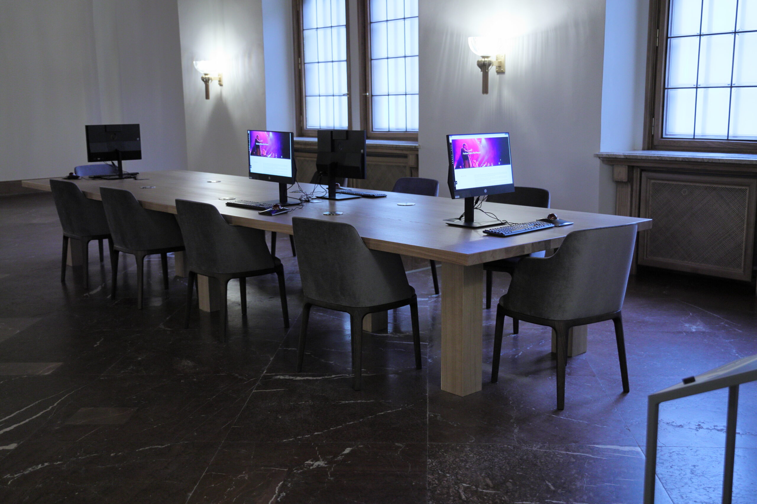 Widok pomieszczenia mediateki: na marmurowej podłodze stoi duży, jasny stół otoczony szarymi krzesłami. Na stole widać cztery monitory, z których dwa są włączone i mają kolorowe ekrany. Za stołem widać ścianę z lampami - kinkietami, z których pada światło, oraz z oknami.