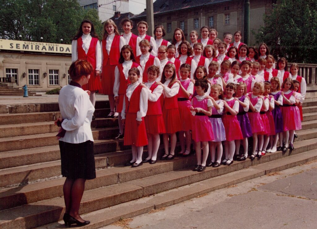 Na kamiennych schodach przed budynkiem Arkadii w Poznaniu stoi grupa dziewcząt w czerwonych spódniczkach, białych bluzkach i czerwonych kamizelkach. W pierwszym rzędzie znajdują się młodsze dziewczynki w kolorowych sukienkach. Wszystkie patrzą na kobietę w czarno-białym kostiumie stojącą przodem do nich, a tyłem do obiektywu. Kobieta wykonuje gest dyrygenta.