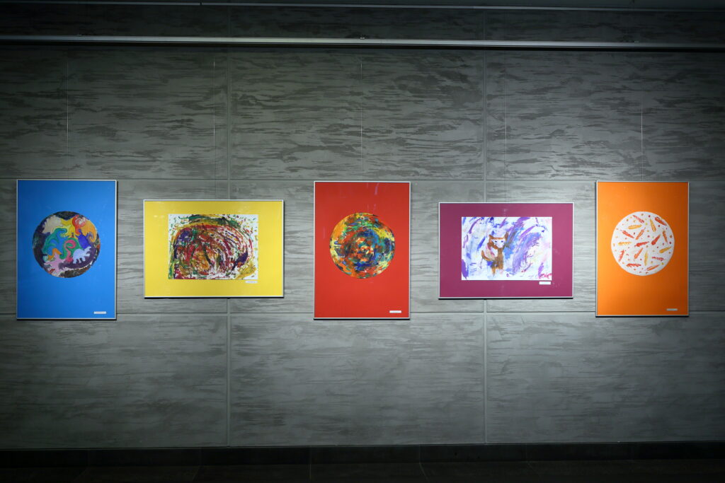 Na ścianie wisi pięć prac uczestników Pracowni. Trzy pionowe z namalowanymi kolorowymi kołami oraz dwie poziome – jedna przedstawia barwną abstrakcję, a druga psa.