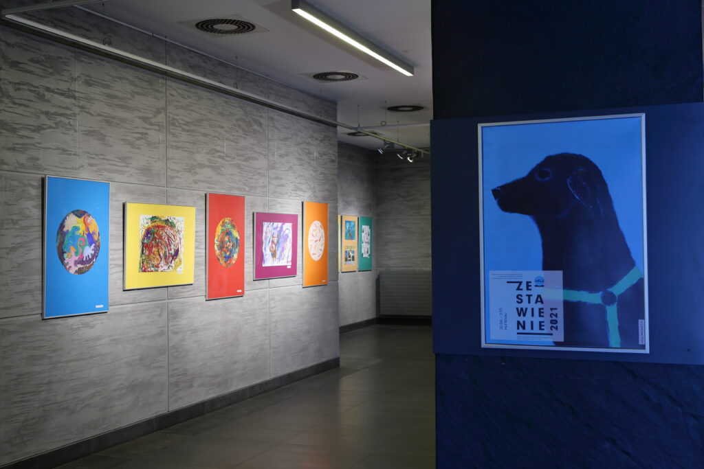 Po lewej stronie zdjęcia widać pięć prac uczestników Pracowni. Trzy pionowe z namalowanymi kolorowymi kołami oraz dwie poziome – jedna przedstawia barwną abstrakcję, a druga psa. W oddali można dostrzec kolejne dwie prace, ale nie widać, co jest na nich namalowane. Po prawej stronie zdjęcia znajduje się plakat promujący wystawę z napisem: „Zestawienie 2021”.