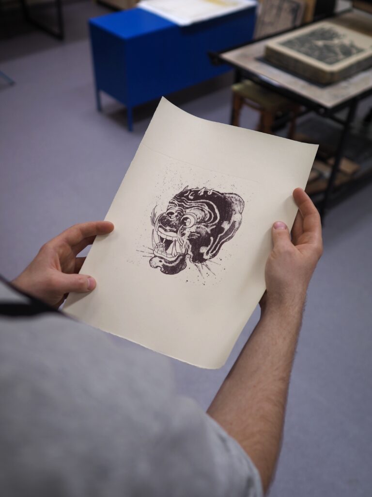 Na zdjęciu litografia trzymana w rękach niewidocznego mężczyzny. Praca przedstawia głowę zwierzęcia lub postaci mitycznej. Zdjęcie wykonana z góry. W tle widoczna szara podłoga, a w oddali dwa stoły – jeden niebieski, drugi czarny.