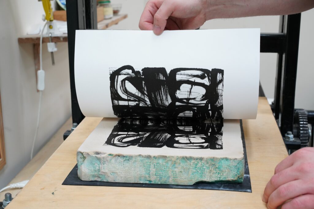 Na stole prasy litograficznej leży kamień z przygotowaną litografią. Odbywa się proces odbijania grafiki – widać rękę, która zdejmuje z matrycy odbitkę graficzną.