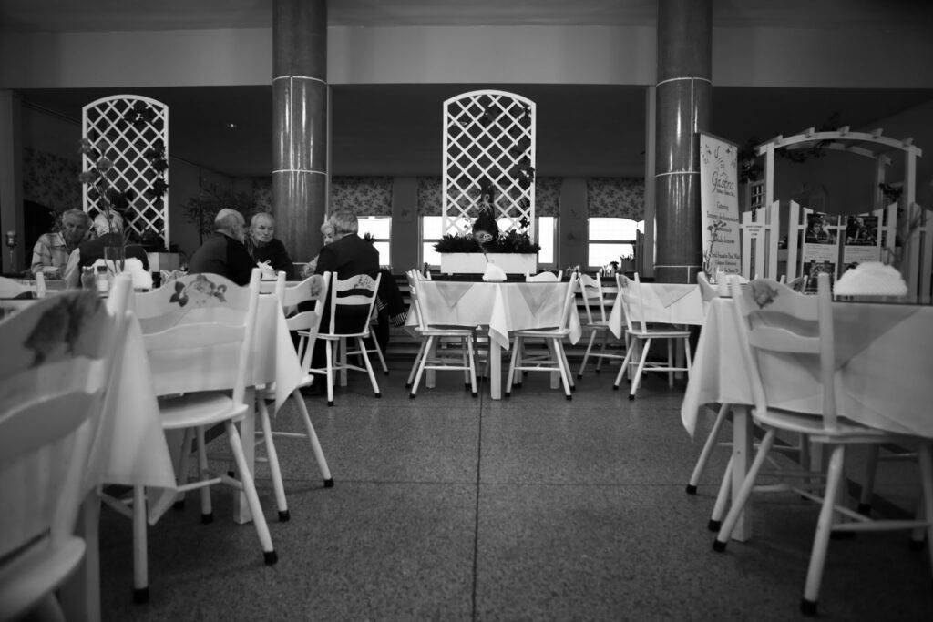 Fotografia czarno-biała. Wnętrze kawiarni, zdjęcie robione „z biodra”, nisko. Po prawej i lewej stronie białe stoliki i krzesła. W głębi wyostrzony kadr, dwie kolumny podpierają sufit, widzimy cztery stoliki, dwa po lewej są zajęte przez dwie grupki starszych osób. Wzrok przyciągają dwa białe, ażurowe panele, na których wiszą doniczki z kwiatami. Za nimi białymi prostokątami świecą okna.