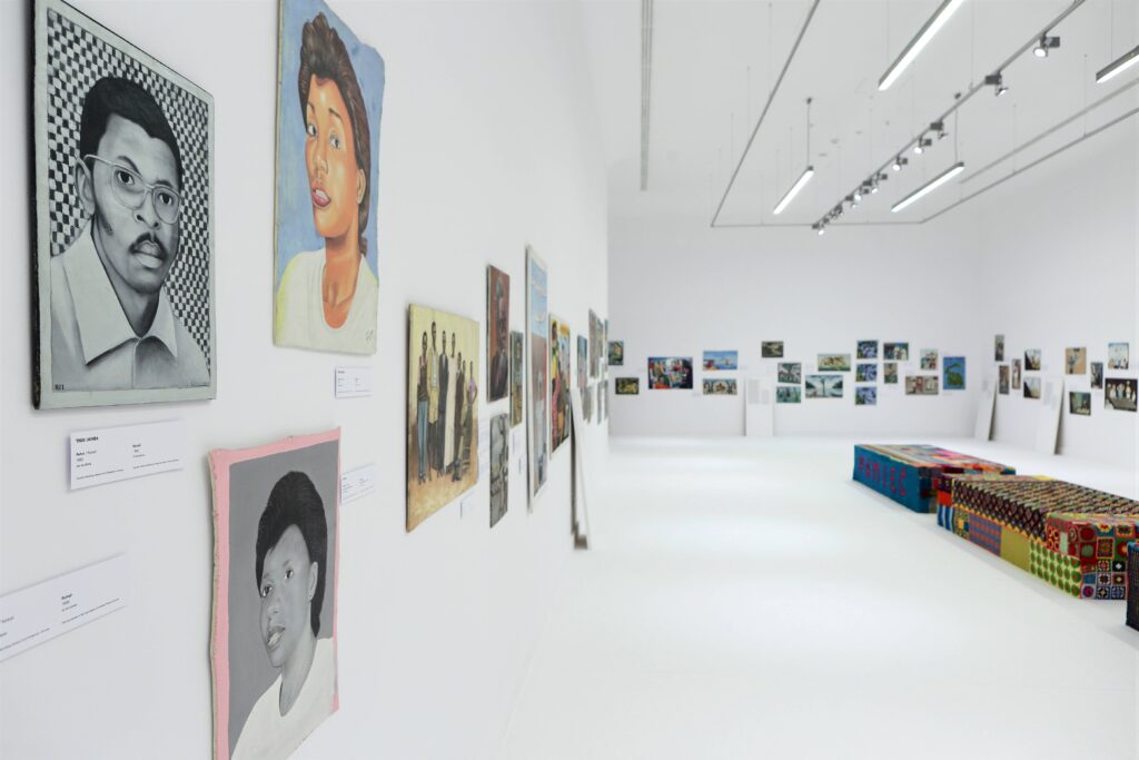 Zdjęcie przedstawia białą salę galerii z obrazami rozmieszczonymi wzdłuż wszystkich ścian. Na fotografii widoczna jest również instalacja złożona z kolorowych skrzyń umieszczonych na podłodze. Po lewej stronie zdjęcia mocne zbliżenie na portrety dwóch kobiet i mężczyzny.
