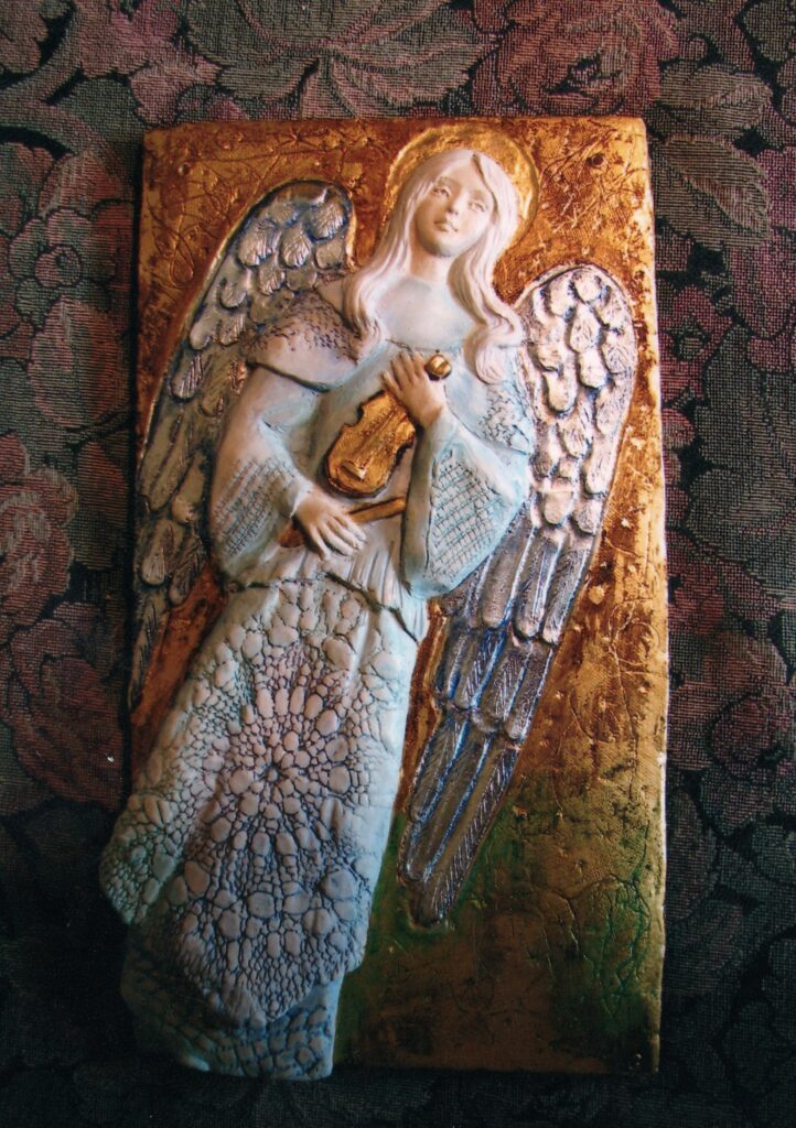Prostokątna płaskorzeźba przedstawiająca anioła ze skrzypcami. Tło rzeźby, aureola, skrzypce i smyczek są pozłacane i mienią się w kontraście do matowego, białego anioła. Jego szata jest bogato zdobiona, z detalami na dole sukni i rękawach. Na skrzydłach wyrzeźbiono małe pióra. Również twarz i włosy są pełne detali, a mina anioła łagodna.