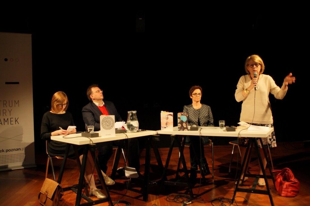 Klucze do Wyobraźni: Inga Iwasiów / Fot. Maciej Kaczyński, CK ZAMEK / Cztery osoby biorące udział w rozmowie z Ingą Iwasiów. Siedzą przy stole – białym blacie na czarnych koziołkach,  na którym stoją książki, szklanki, dzbanek, leżą mikrofony. Od lewej siedzą: prowadząca dyskusję Lucyna Marzec – ma blond włosy do ramion, ciemne oprawki okularów, czarną sukienkę, spogląda w dół, notuje. Obok prowadzącej Krzysztof Uniłowski w ciemnej marynarce, czerwonym swetrze, spod którego wystaje kołnierzyk białej koszuli. Uniłowski ma krótkie brązowe włosy, okulary w jasnych oprawkach. Dalej – Inga Iwasiów: patrzy przed siebie, ma krótkie ciemne włosy, okulary w ciemnych oprawkach, marynarkę bez kołnierzyka w szaro-czarny geometryczny wzór. Na skraju, po prawej stronie Bogumiła Kaniewska w jasnym swetrze, wyrazistym naszyjniku, spódnicy w kolorowy wzór. Kaniewska ma półdługie jasne włosy, okulary w ciemnych oprawkach, stoi, mówi do mikrofonu, który trzyma w prawej ręce, lewą gestykuluje.