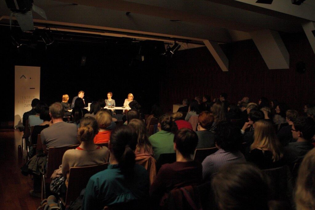 Klucze do Wyobraźni: Inga Iwasiów / Fot. Maciej Kaczyński, CK ZAMEK / Ciemne wnętrze sali, zdjęcie zrobione z ostatnich rzędów, zza pleców licznie zgromadzonej publiczności. Z przodu, w jasnej plamie światła, widzimy panelistów: trzy kobiety i mężczyznę, siedzących przy stole. Są nieco przysłonięci przez głowy osób siedzących na widowni. Z lewej strony wyróżnia się jasny, prostokątny roll-up Centrum Kultury ZAMEK.