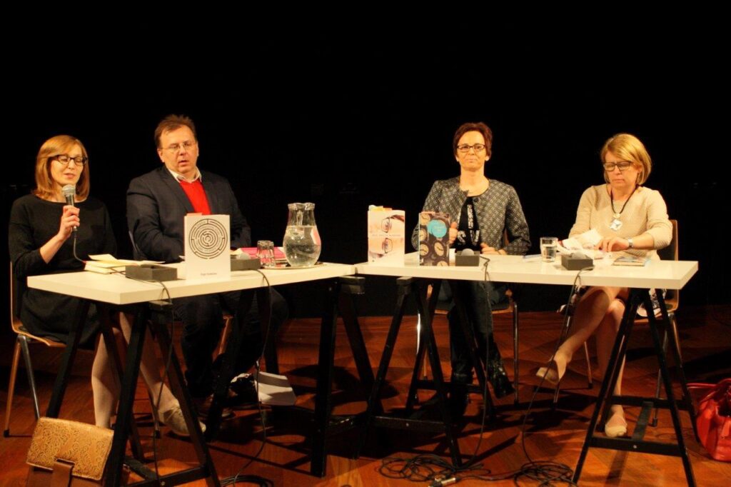 Klucze do Wyobraźni: Inga Iwasiów / Fot. Maciej Kaczyński, CK ZAMEK / Cztery osoby biorące udział w rozmowie z Ingą Iwasiów. Siedzą przy stole – białym blacie na czarnych koziołkach – na którym stoją książki, szklanki, dzbanek, leżą mikrofony. Od lewej siedzą: prowadząca dyskusję Lucyna Marzec: ma blond włosy do ramion, ciemne oprawki okularów, czarną sukienkę, trzyma mikrofon, do którego mówi. Obok prowadzącej: Krzysztof Uniłowski w ciemnej marynarce, czerwonym swetrze, spod którego wystaje kołnierzyk białej koszuli. Uniłowski ma krótkie brązowe włosy, okulary w jasnych oprawkach. Dalej – Inga Iwasiów: patrzy przed siebie, ma krótkie ciemne włosy, okulary w ciemnych oprawkach, marynarkę bez kołnierzyka w szaro-czarny geometryczny wzór. Na skraju, po prawej stronie Bogumiła Kaniewska w jasnym swetrze, wyrazistym naszyjniku. Kaniewska ma półdługie jasne włosy, okulary w ciemnych oprawkach; patrzy w dół, przegląda notatki.
