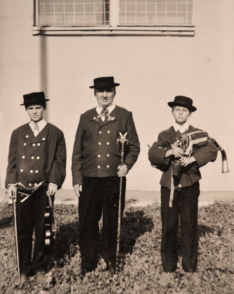 Trzech muzyków w strojach ludowych, jeden ze skrzypcami, dwóch z dudami pozują do fotografii na tle budynku.