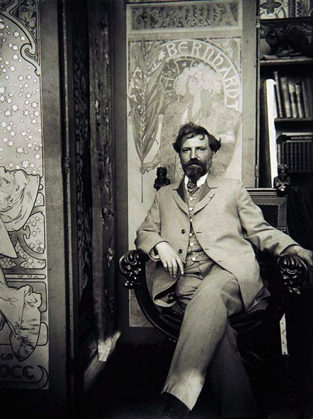 Fotografia jest czarno-biała, przedstawia Alfonsa Muchę w swojej pracowni. Mężczyzna siedzi na rzeźbionym krześle lub fotelu, jego sylwetka umiejscowiona została w lewej, dolnej części fotografii. Mężczyzna ma na twarzy zarost – bujne wąsy oraz brodę. Spogląda w obiektyw. Lewą rękę trzyma na oparciu siedziska, prawym łokciem podpiera drugie oparcie. Ma założoną lewą nogę na prawą. Ubrany jest jasny strój.  Po prawej stronie fotografii widać fragment regału z książkami. W centrum oraz po lewej stronie tła przedstawione są prace artysty – w centrum jest to Gismonda; praca całkiem po lewej jest jedynie fragmentem.