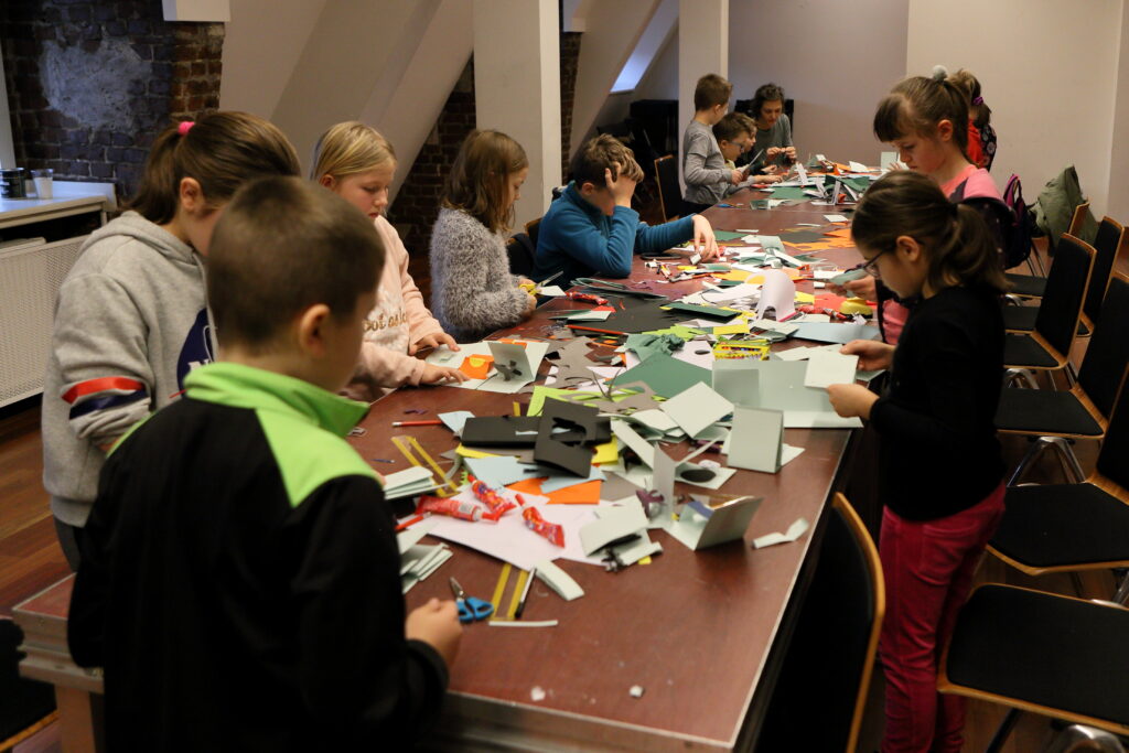 Długi stół, na którym leży mnóstwo arkuszy kolorowego papieru. Właśnie z niego, znajdujące się przy stole dzieci, wycinają kolorowe wzory. W oddali, na końcu stołu instruktorka pracuje razem z młodymi twórcami.