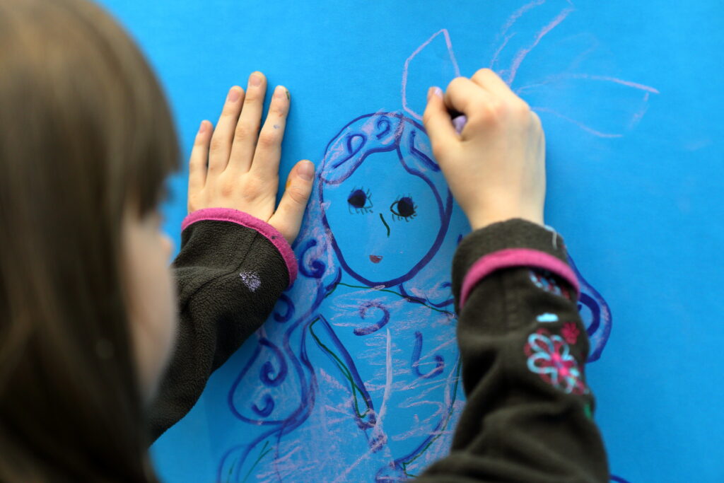 Dziecko, zwrócone tyłem do obiektywu, rysuje na niebieskiej kartce kobietę z długimi włosami. Lewą ręką opiera się o swoje dzieło.