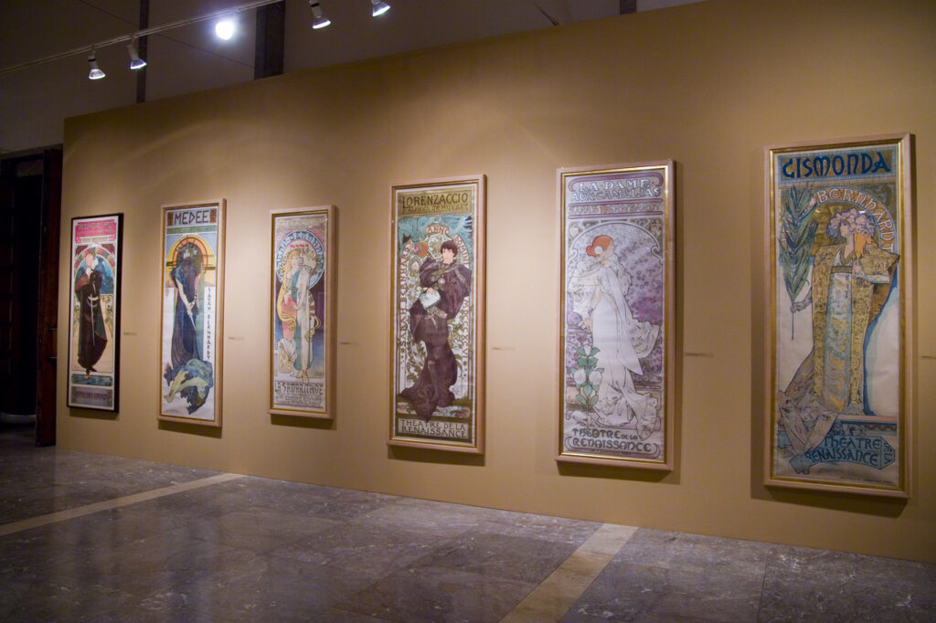Fotografia przedstawia fragment Sali Wystaw. W jej wnętrzu na jasnobeżowej ścianie wiszą duże kolorowe plakaty z okresu secesji. To portrety kobiet ubranych w zwiewne suknie. W tle obrazów widać kwiatowe motywy.