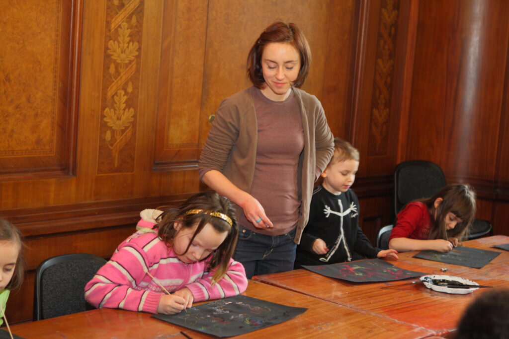 Czworo dzieci rysuje na czarnych kartkach. Ich pracom przygląda się instruktorka.