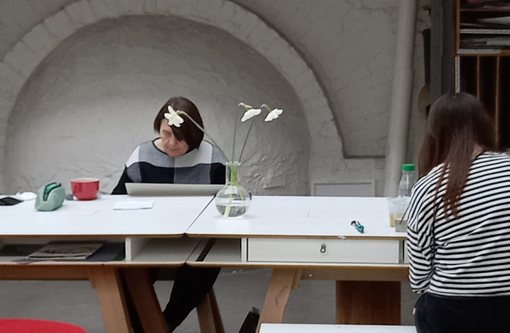 Fotografia wykonana w pracowni plastycznej. Przy długim białym stole, na którym stoi wazon z narcyzami, pracują dwie uczestniczki warsztatów.
