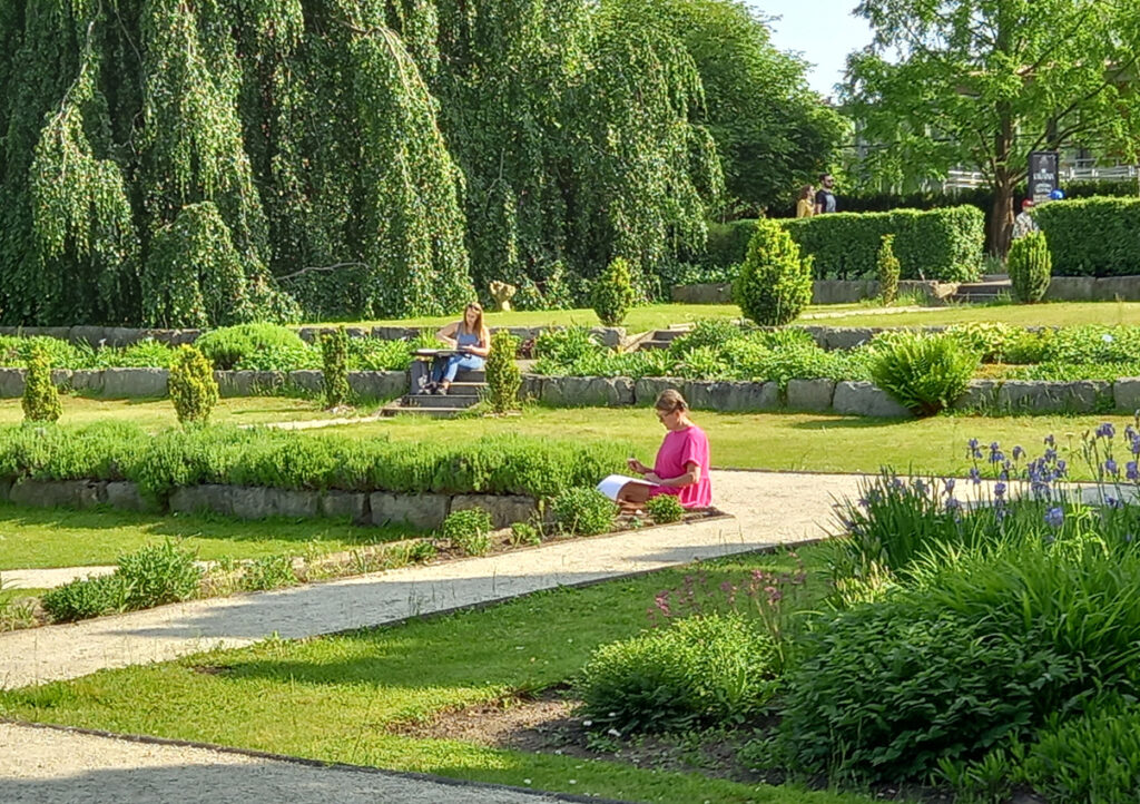 Plener malarski w Ogrodzie Botanicznym. W przestrzeni parku, gdzie dominują zieleń traw i drzew, dwie uczestniczki warsztatów siedzą na schodkach i malują z natury.