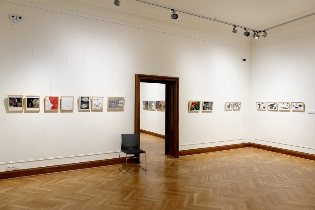 Po lewej stronie zdjęcia na ścianie umieszczono prace uczestników i uczestniczek warsztatów malarskich. W centrum fotografii otwarte drzwi do drugiego pomieszczenia z kolejnymi pracami z wystawy. Po prawej stronie kolaże.