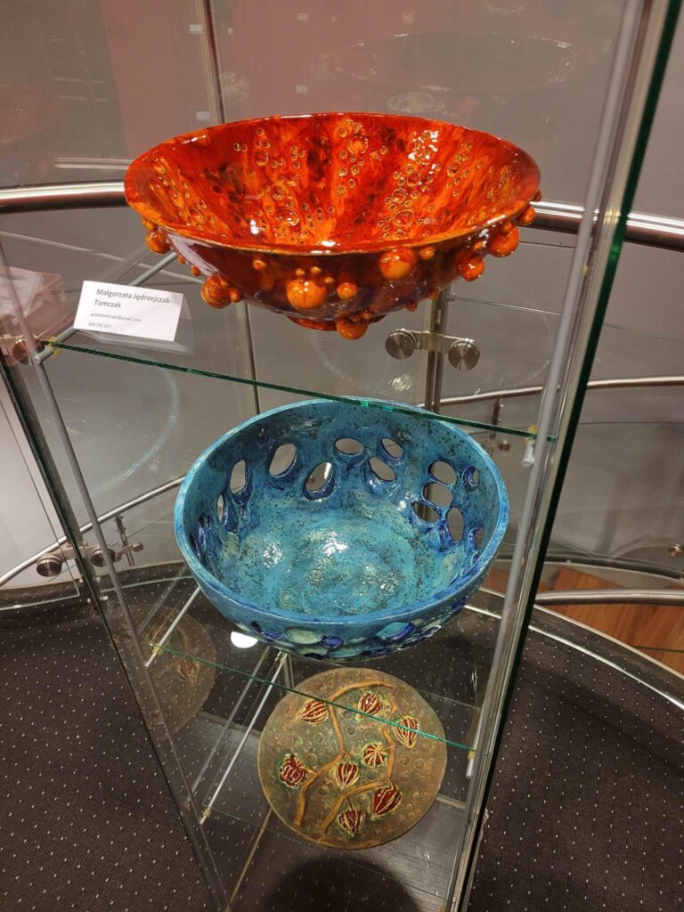 Zbliżenie na szklaną gablotę, w której znajdują się trzy prace ceramiczne: misy w czerwonym, niebieskim i brązowym kolorze. W tle, za szklaną szybą, widoczne są schody.