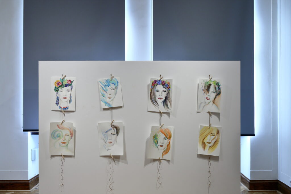 Na pierwszym planie, na białej ściance osiem małych portretów kobiet w pastelowej kolorystyce na papierze. W tle okna z zasłoniętymi roletami.