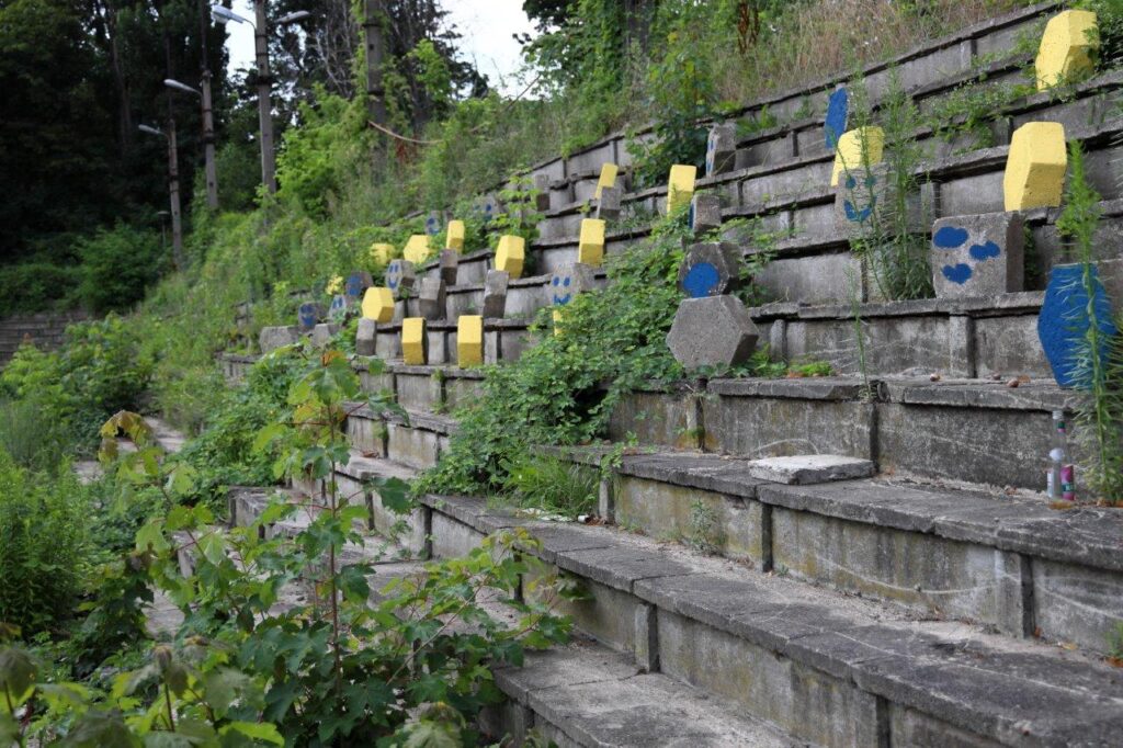 Fragment starej, betonowej trybuny stadionowej, poprzerastanej roślinami. Na schodkach stoją pomalowane na żółto kostki betonowe.