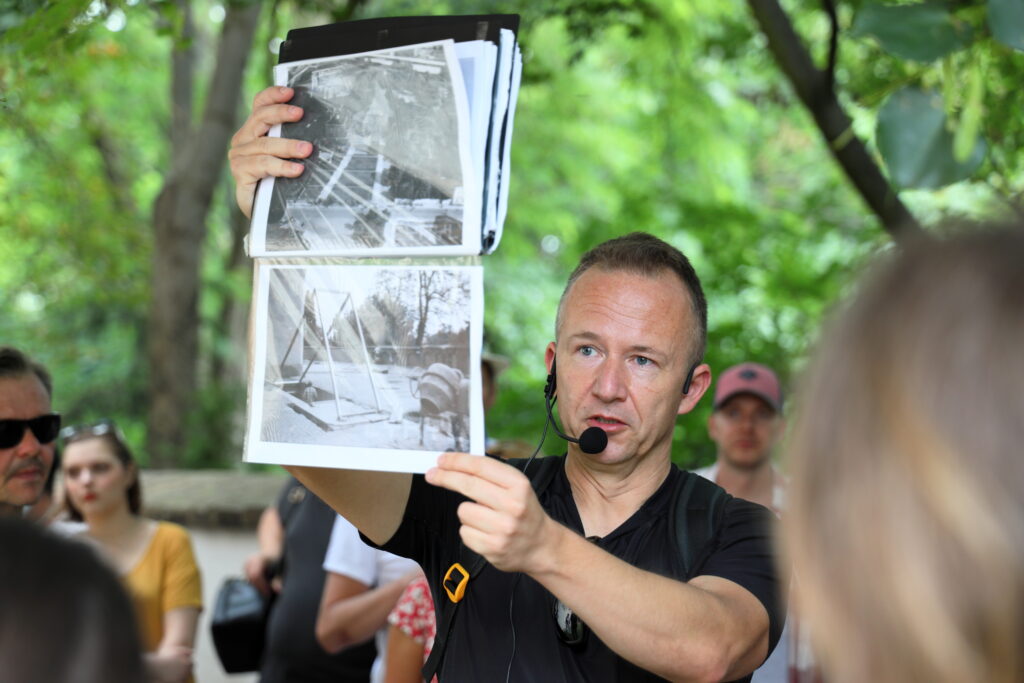 Osoba męska z założonym mikrofonem stoi wśród ludzi i drzew, w wyciągniętych wysoko rękach trzyma skoroszyt z czarno-białymi fotografiami