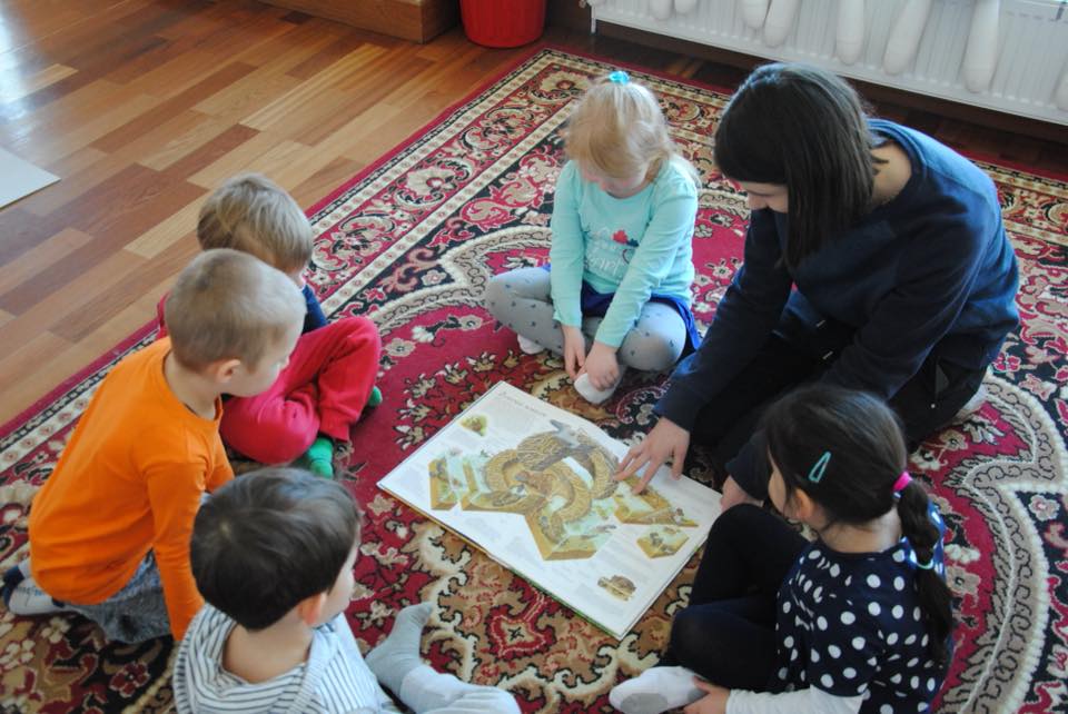 Na dywanie, wraz z panią prowadząca zajęcia, siedzą dzieci po turecku. Przed nimi rozłożona jest plansza z rysunkiem budowli. Dzieci patrzą na planszę.
