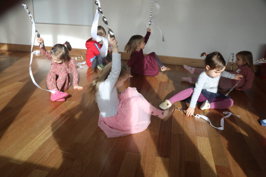 Sześcioro dzieci siedzących na podłodze, trzyma w uniesionych do góry rękach wstążki gimnastyczne.