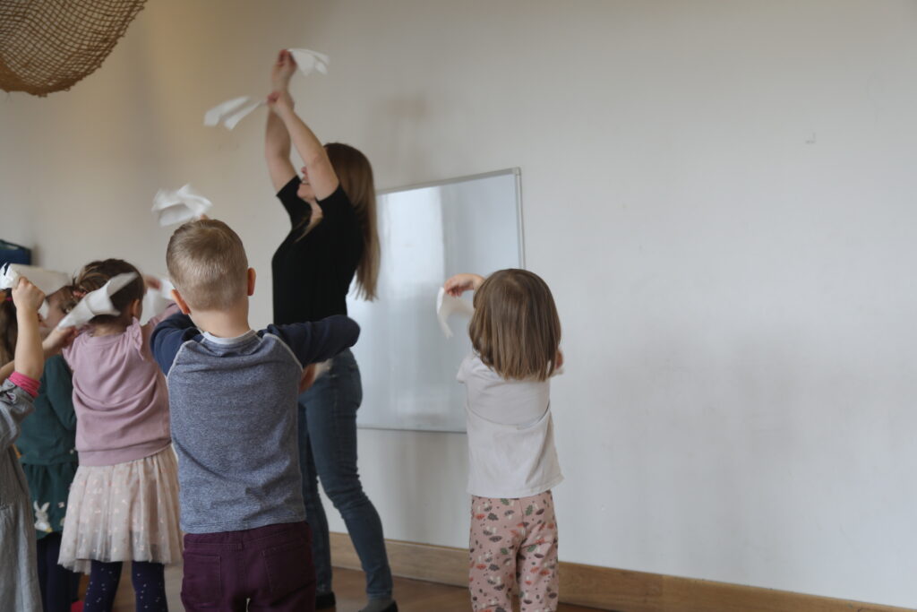Grupa dzieci stoi tyłem do obiektywu. Naśladują ruchy prowadzącej zajęcia – trzymają w obu dłoniach białe chusteczki i machają nimi w różne strony.