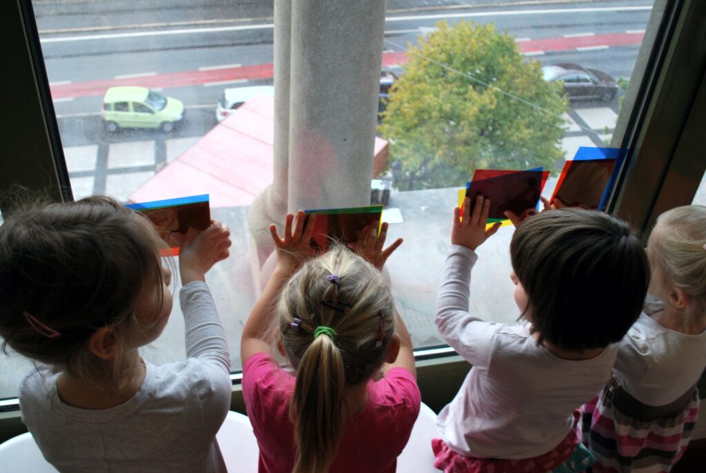 Czwórka dzieci stoi przy oknie tyłem do obiektywu, przykładają do szyby różnego koloru folię, dzięki czemu powstały różne kolory. Za oknem, na ulicy widać samochody i drzewo.