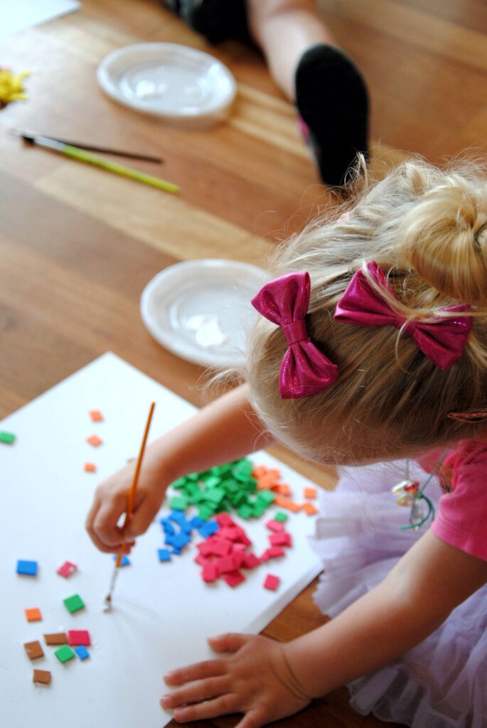 Na podłodze siedzi dziewczynka z dwoma różowymi kokardami we włosach. Pochyla się nad kartką papieru, na którą nanosi pędzelkiem klej. Na kartce rozsypane są kolorowe kawałki mozaiki. W tle fotografii widoczne jest inne dziecko i dwa talerzyki z klejem.