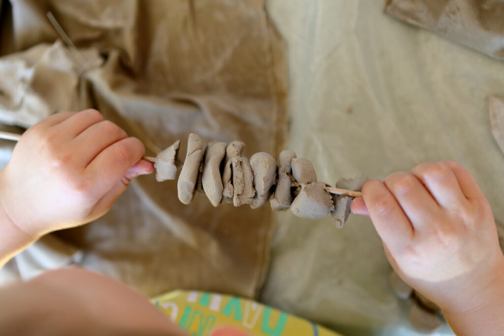 Fotograficzne zbliżenie na ręce dziecka. Trzyma ono drewniany patyk, na który wbite są kawałki gliny.