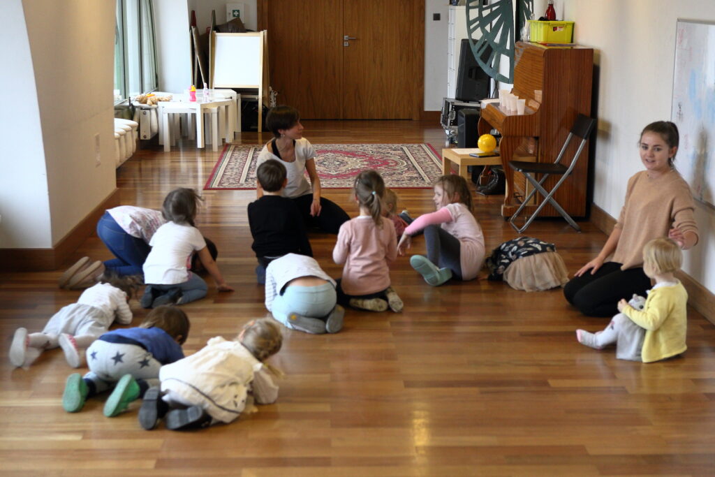 Dzieci siedzą na dywanie, pochylając ciała do podłogi. Na sali obecne są dwie prowadzące – jedna zwrócona jest przodem do obiektywu, a druga bokiem do grupy. W głębi sali widać pianino, krzesło, stoliki, tablicę i dywan. W oddali znajdują się drzwi.