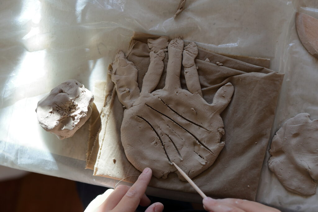 Na stole leży płaskorzeźba dłoni wykonana z gliny. Obok niej dwa dodatkowe kawałki gliny. Widać też dwie dziecięce ręce, z których jedna patykiem rysuje na glinianej dłoni linie papilarne.