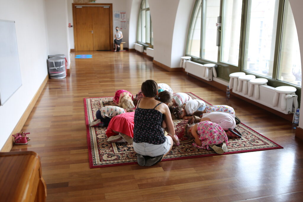 Panorama dużej sali, w której na prawej ścianie są dwa olbrzymie okna, a na tylnej drzwi. Na dywanie w przedniej części sali siedzą dzieci w okręgu. Pochylają się do przodu, tak że głowami dotykają podłogi. Dzieci są pod opieką pań prowadzących zajęcia.