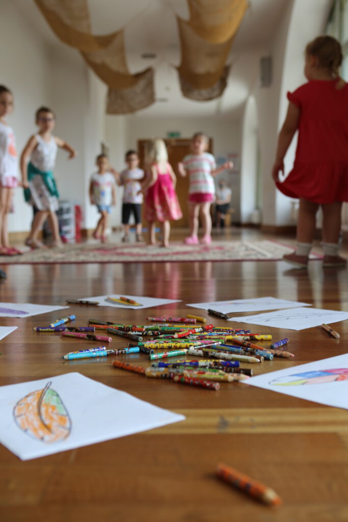 Na pierwszym planie widać rozłożone na podłodze dziecięce rysunki oraz kredki. Na drugim planie, w oddali tańczy grupa dzieci.