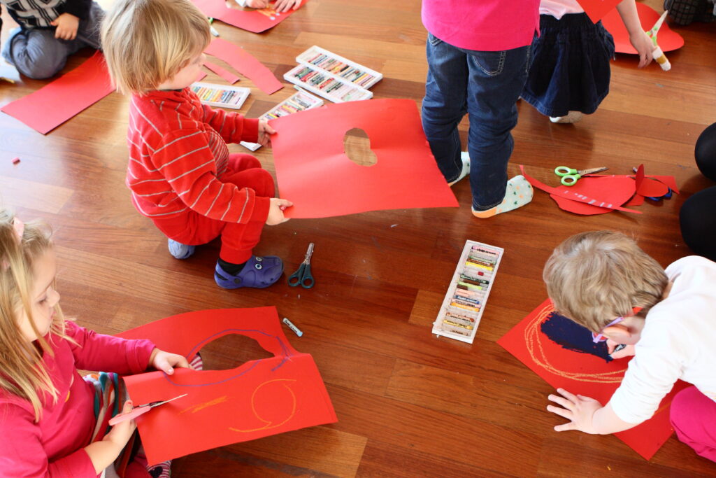 Grupa dzieci wykonuje zadanie plastyczne na podłodze. Rysują na czerwonych kartkach papieru i wycinają je. Na podłodze leżą kredki, nożyczki i skrawki czerwonego papieru.
