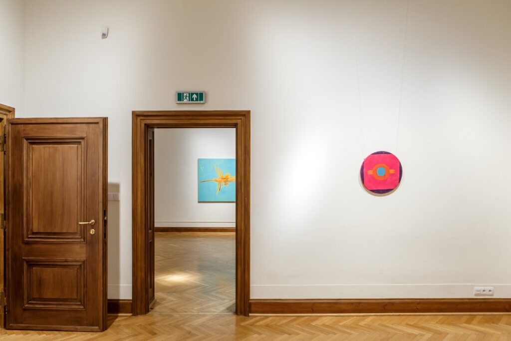 Po prawej strponie obraz w kształcie koła w dominującej czerwieni. W głębi w otwartych drzwiach fragment błękitnego obrazu