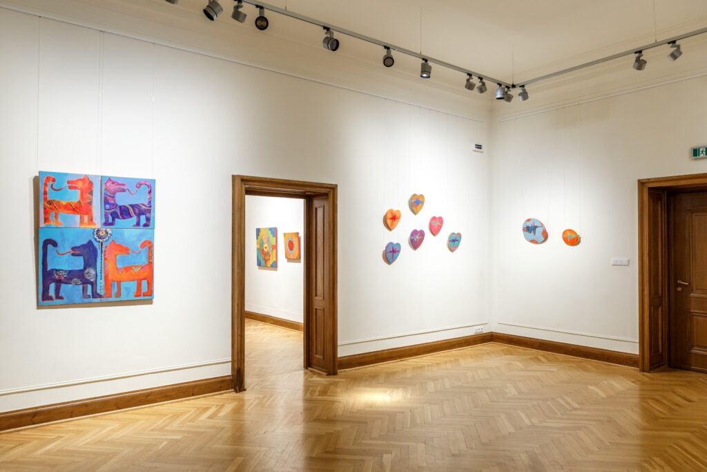 Po lewej stronie obraz z motywem czterych odwróconych smoków. Na wprost kolorowe obrazy z motywem kolibrów. W głębi w otwartych drzwiach dwa kolorowe obrazy